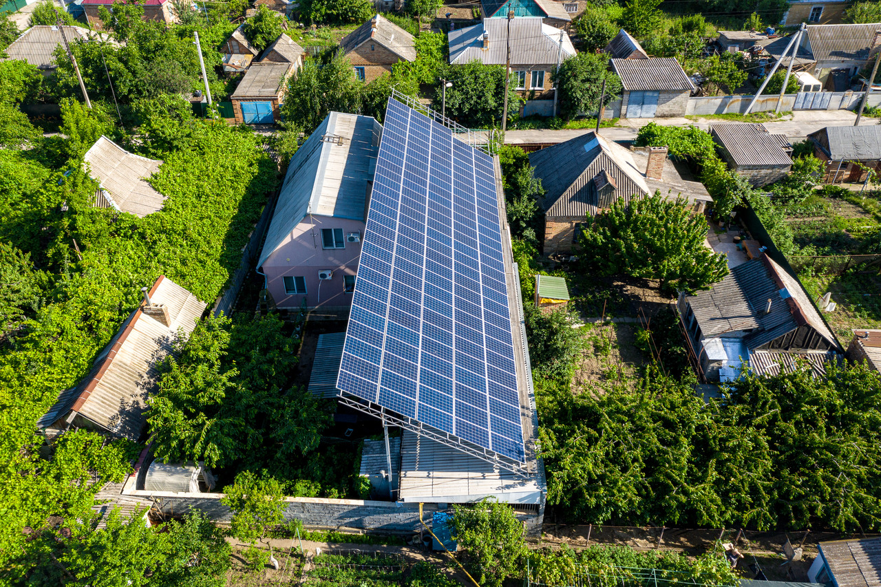 Aerial view of a residential area with one house having a large array of solar panels on its roof, surrounded by green trees and neighboring houses with various roof types.
